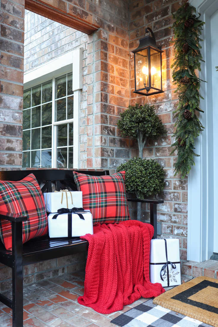 Front porch decor includes bench with plaid pillows red blanket and Christmas presents