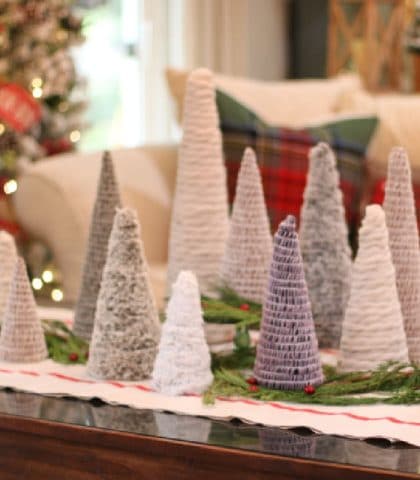mini Christmas trees of different shapes and textures displayed on bed of evergreens with Christmas tree lights in background